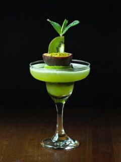 tall margarita glass filled with green liquid, kiwi slice and mint on a black background.