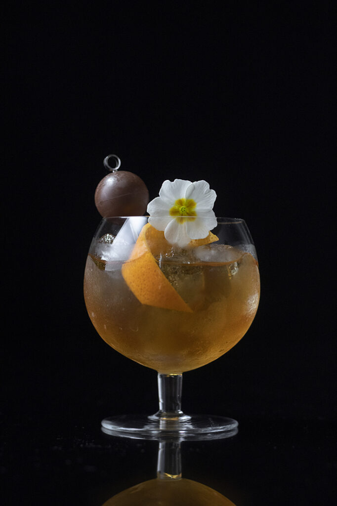 a cognac cocktail in a small snifter with a chocolate truffle, orange twist, and a white flower.