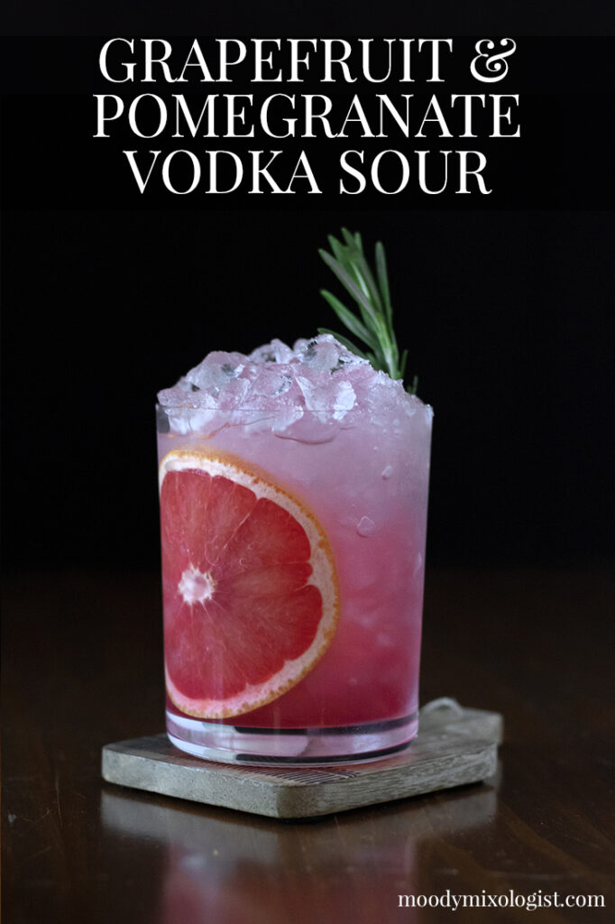 a pink drink in a rocks glass with a red grapefruit wheel and a rosemary sprig.