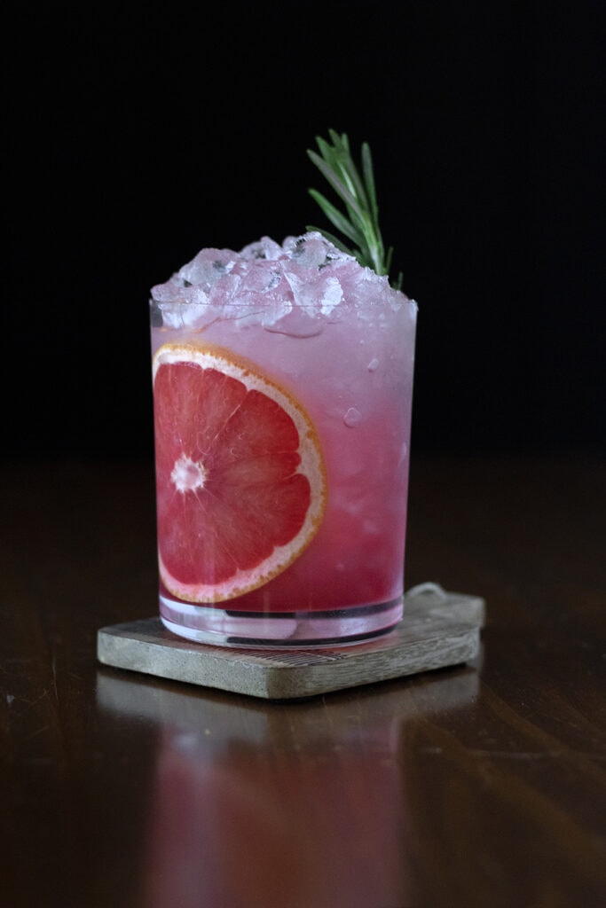 a pink drink in a rocks glass with a red grapefruit wheel and a sprig of rosemary.