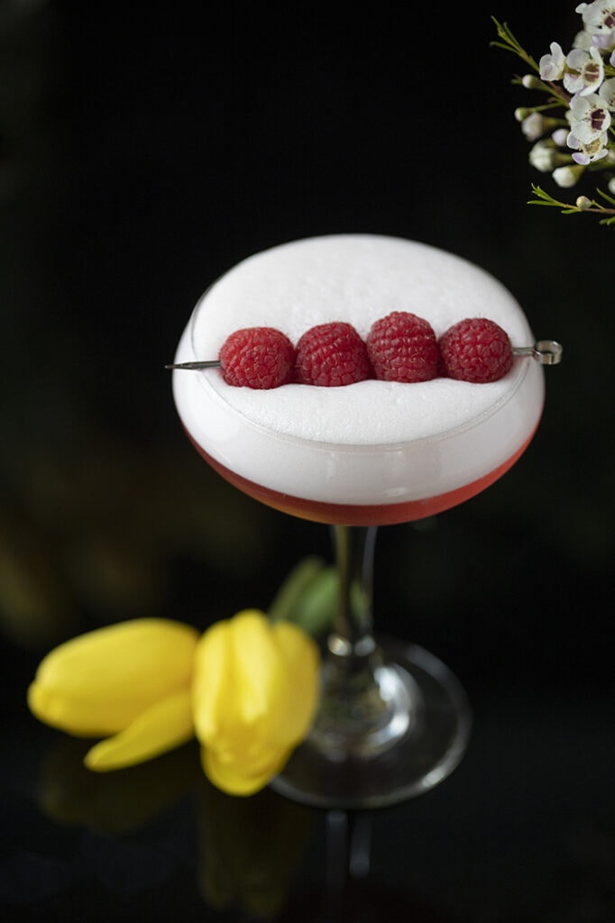 red cocktail with thick white foam on top and raspberries for garnish