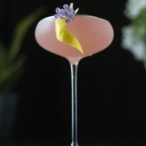 pink cocktail on a black background with a lemon twist and flower