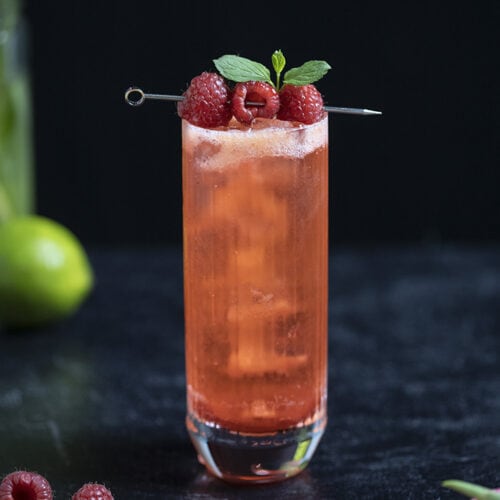highball glass with red clear cocktail garnished with raspberries and mint