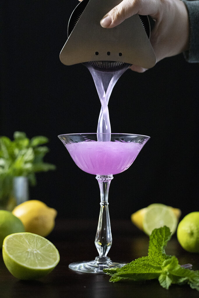 pouring a bright purple cocktail into a coupe glass