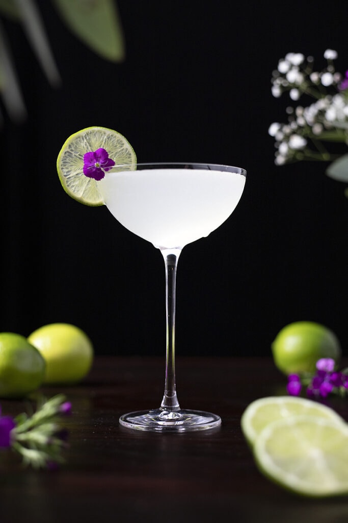 a frosty clear cocktail in a tall coupe glass with a lime wheel and small flower.
