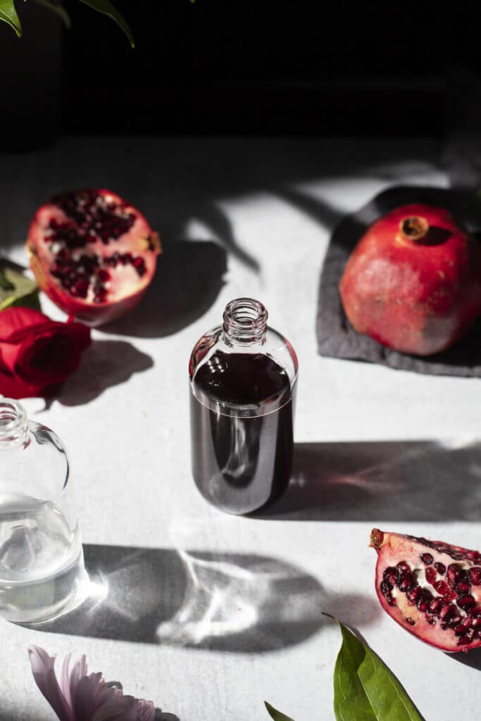 a bottle of dark red syrup surrounded by pomegrantes and flowers