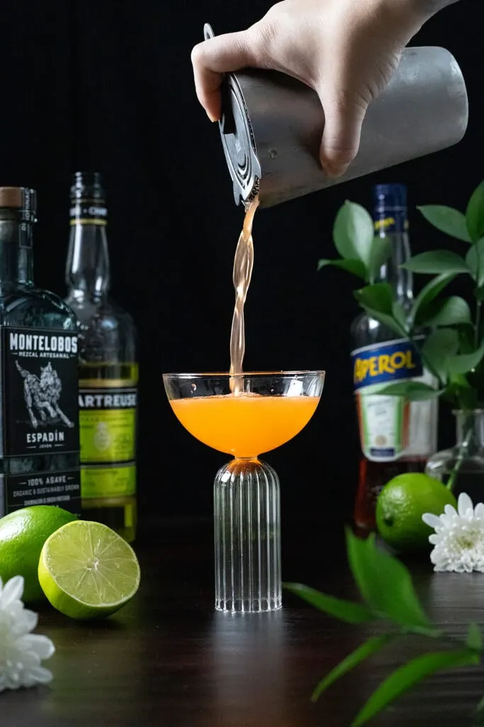 straining an orange cocktail into a coupe glass.