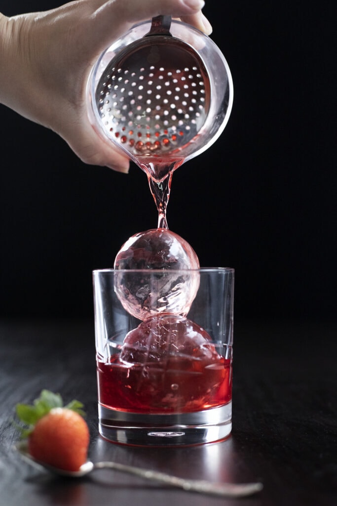 pouring a red cocktail over clear ice balls.