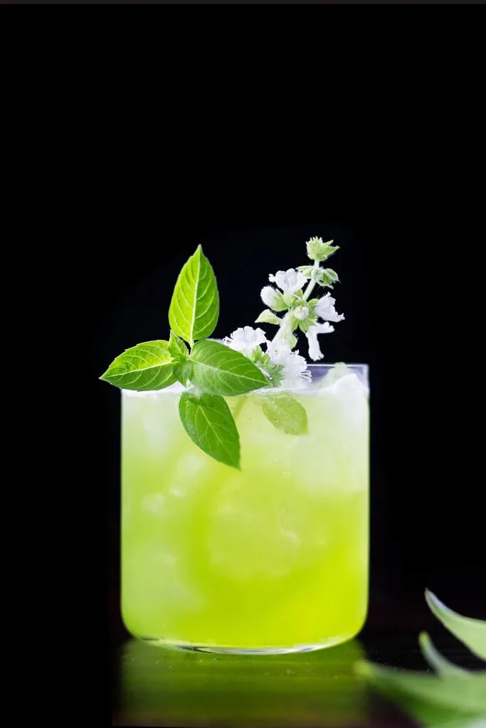 a bright green drink in a rocks glass with a white basil blossom.