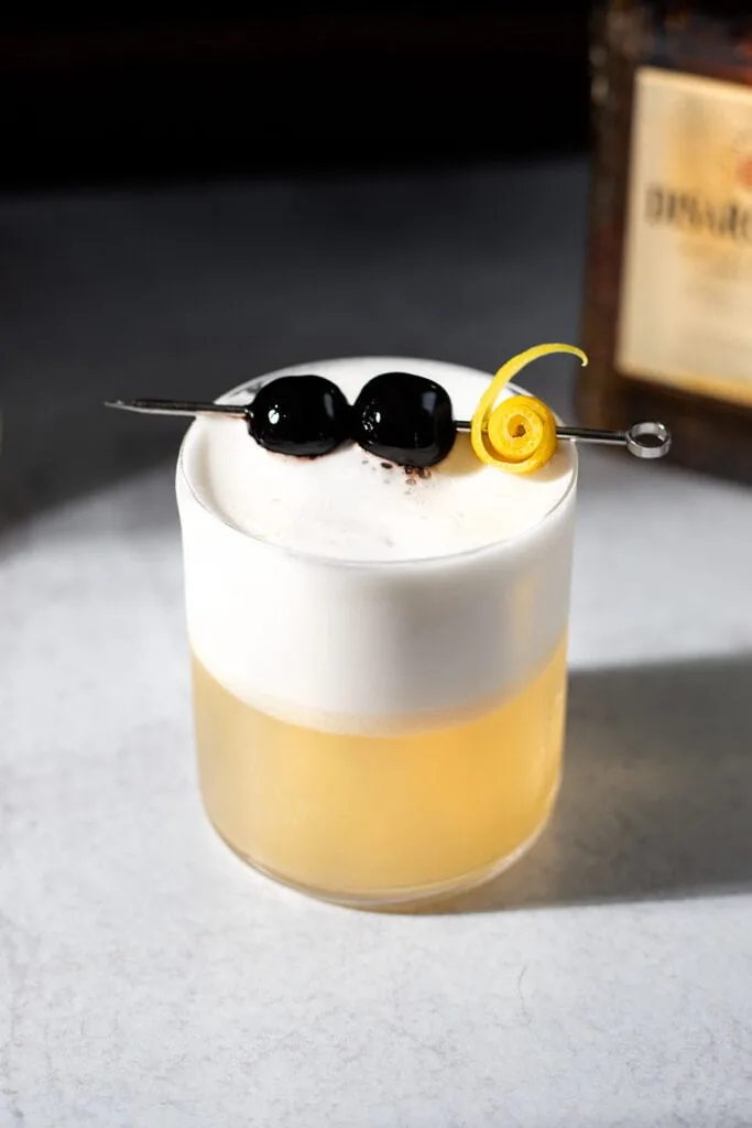 amaretto sour cocktail garnished with two cherries and a fancy lemon twist.