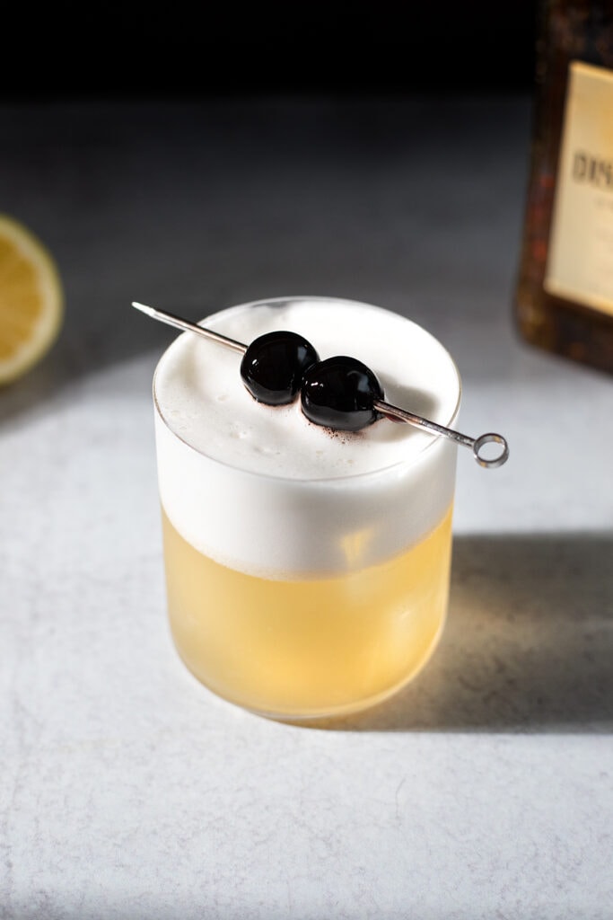 a golden colored cocktail with thick white foam and two cherries.