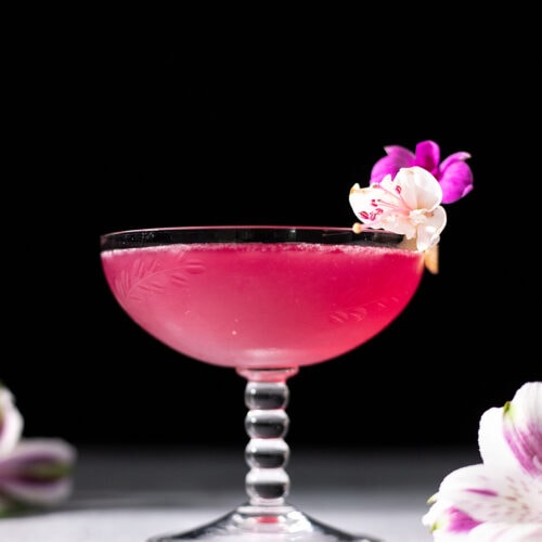 an antique cocktail glass filled with a bright pink liquid and garnished with small flowers.