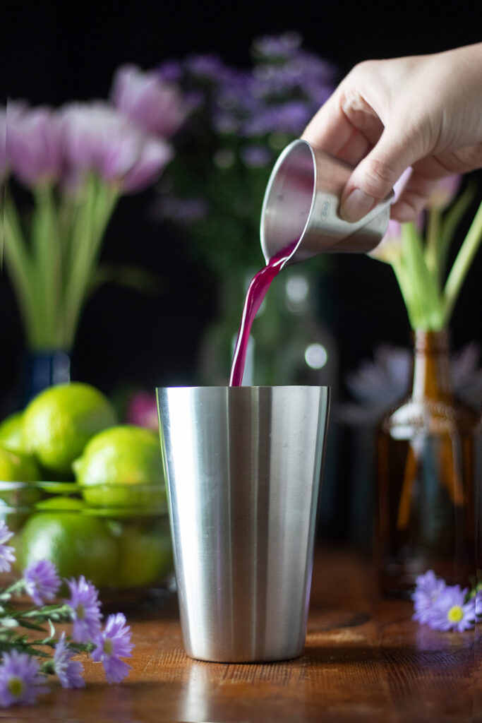 pouring pink syrup from a jigger into a cocktail shaker.