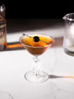 a vintage cocktail glass filled with a brown drink and a cherry on a pick.