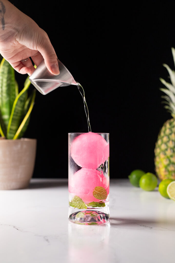 pouring white rum into a glass filled with pink ice spheres.