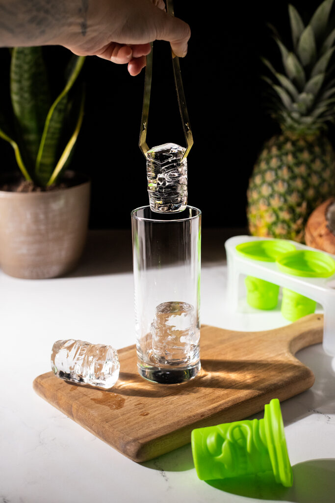 using ice tongs to place clear tiki ice cubes into a tall glass.