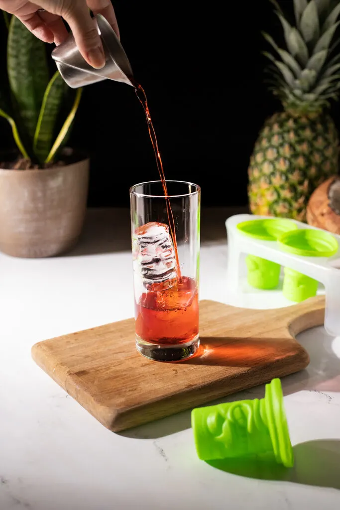 pouring red liqueur over clear tiki ice in a highball glass.