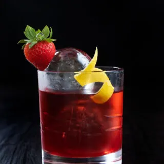 a bright red drink in a rocks glass with a strawberry and a lemon twist.