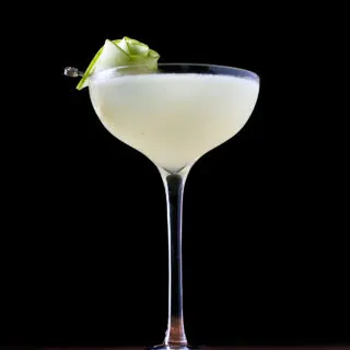 a pale yellow cocktail in a tall coupe glass with a rose garnish made from green apple slices.