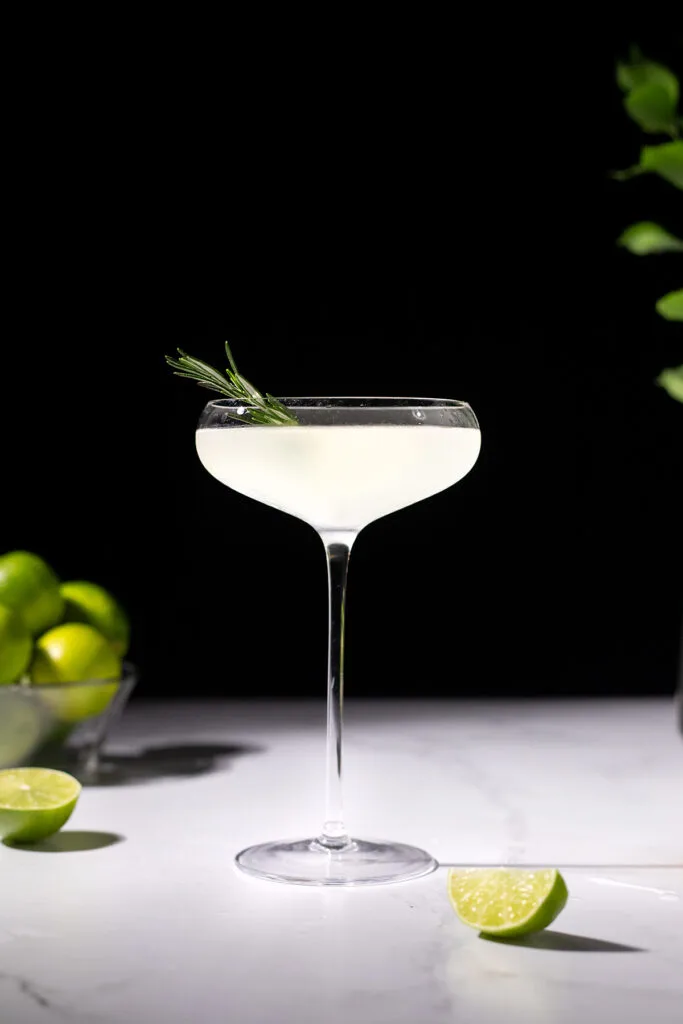 a clear cocktail in a tall coupe glass with a sprig of rosemary.