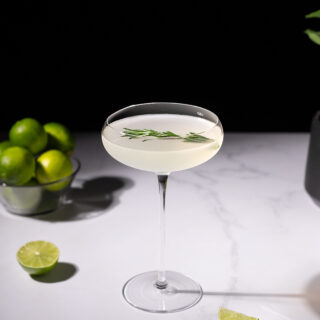 a cocktail in a tall stemmed coupe glass with a rosemary sprig next to sliced limes.