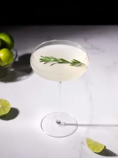 a pale cocktail in a coupe glass next to a bowl of limes.