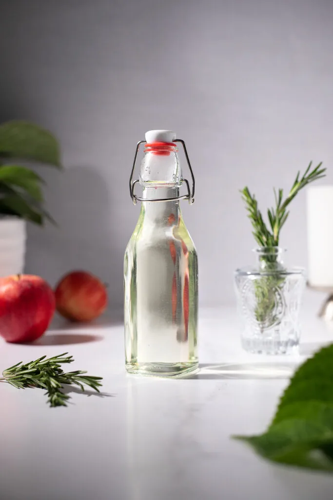 a swing top bottle full of pale green syrup next to rosemary and apples.