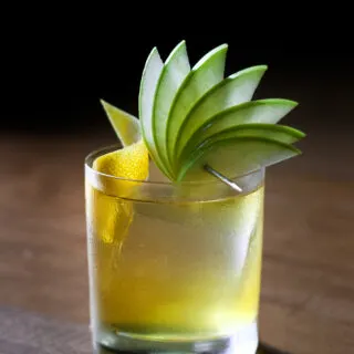 a yellow drink in a rocks glass garnished with a lemon twist and a fan of green apple slices.