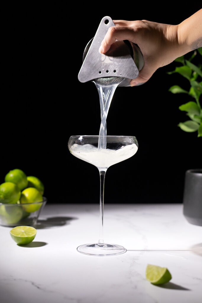 pouring a clear cocktail into a tall coupe glass.
