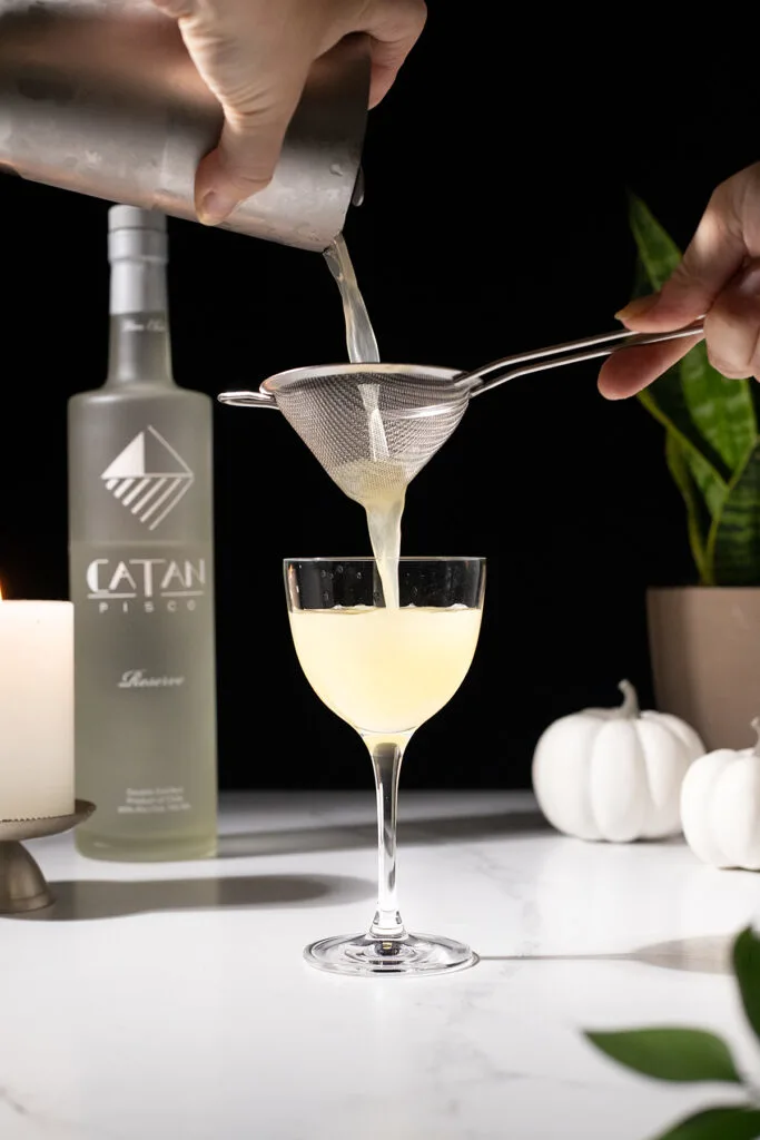 pouring a pale yellow cocktail through a fine mesh strainer into a cocktail glass.