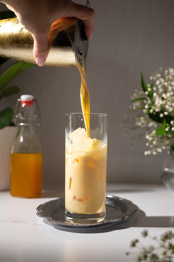 pouring golden milk from a cocktail shaker into a glass filled with ice.