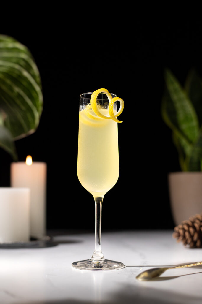 a yellow bubbly cocktail in a champagne flute with a long lemon twist.