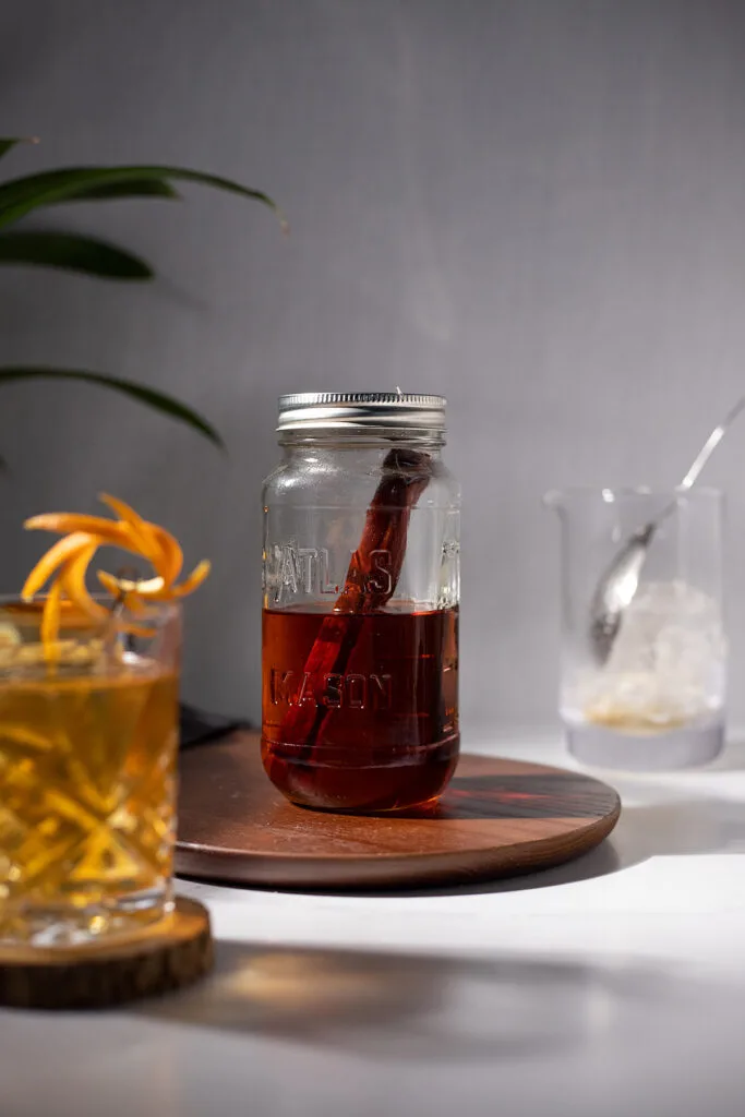 a jar of dark brown liquor with a stick of cherry wood next to a whiskey cocktail and mixing glass.