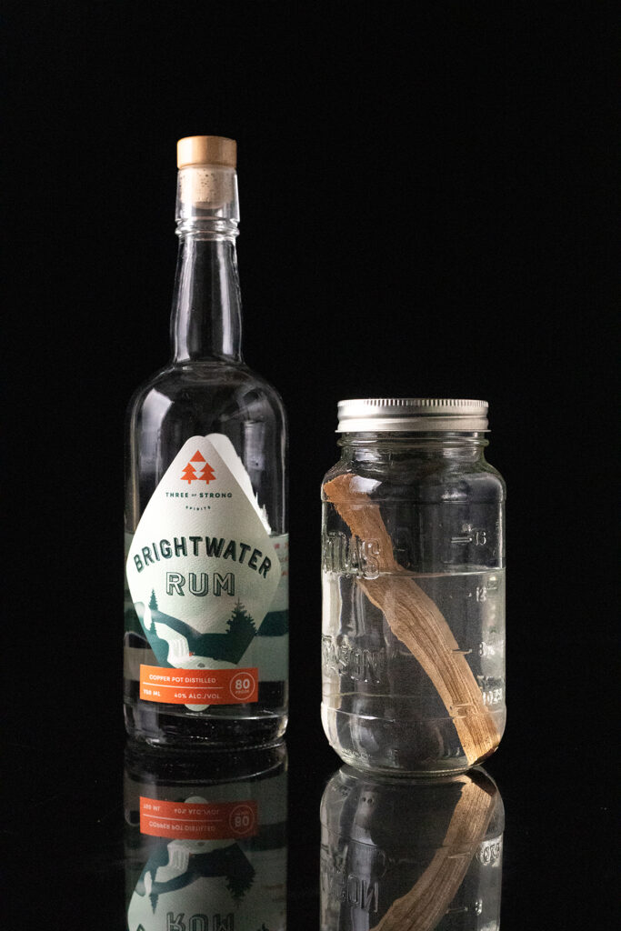 a bottle of Brightwater white rum next to a jar filled with white rum and a stick of maple wood.