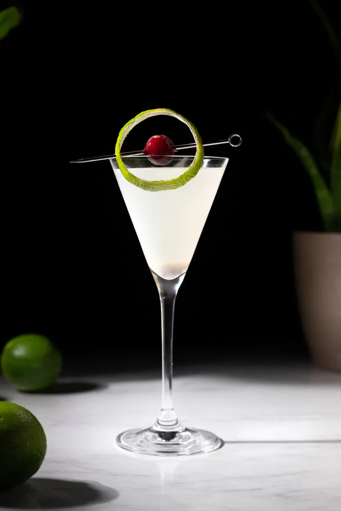 a martini glass filled with a white drink and garnished with a skewered cranberry and lime peel.