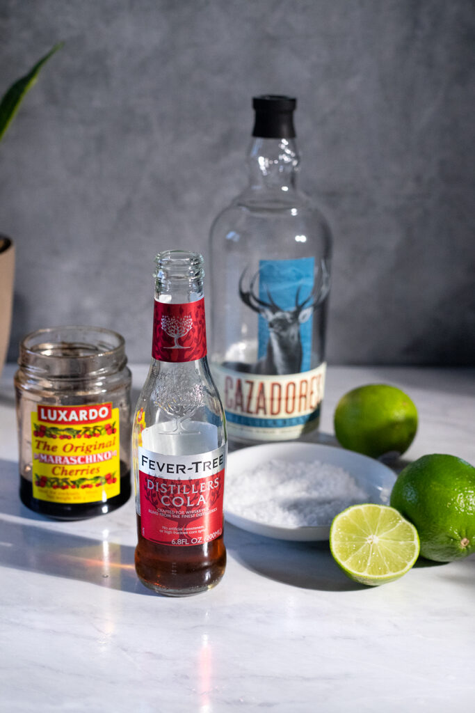 a bottle of tequila with a jar of cherries, a cola bottle, limes and salt.