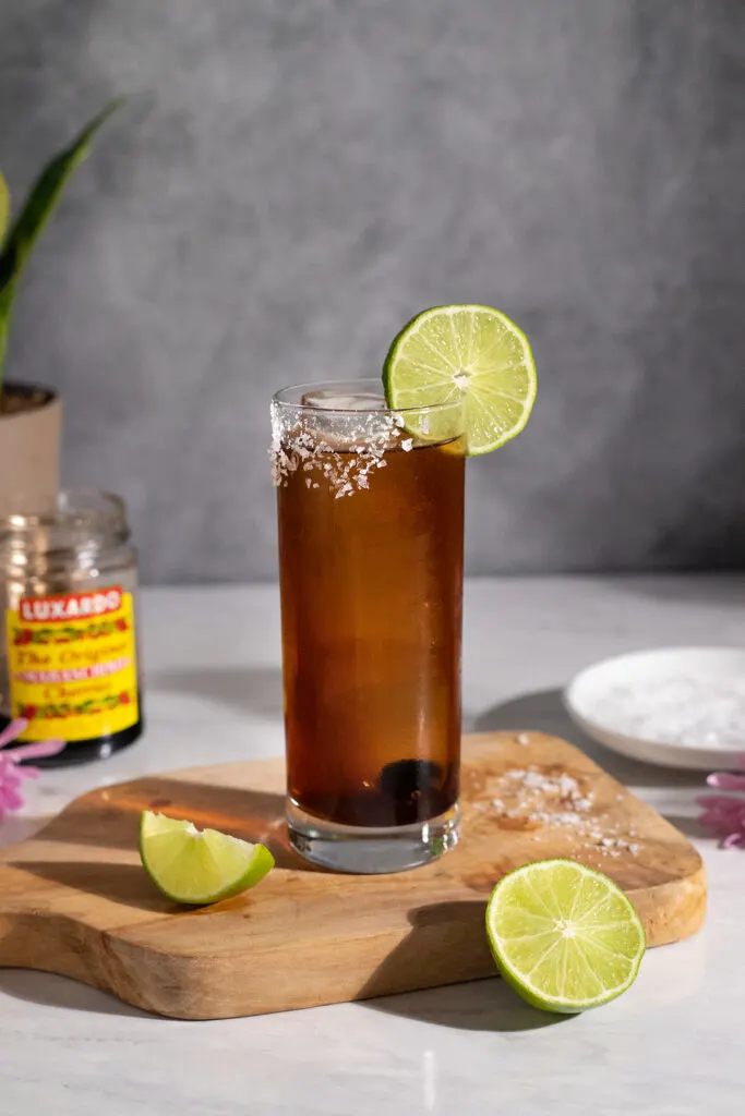 a brown cocktail with a lime wheel on a wooden cutting board.