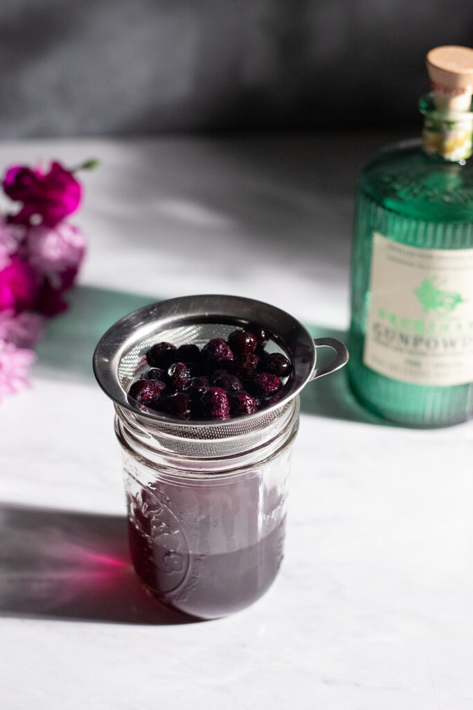 a mason jar with blueberry infused gin and a strainer full of blueberries.