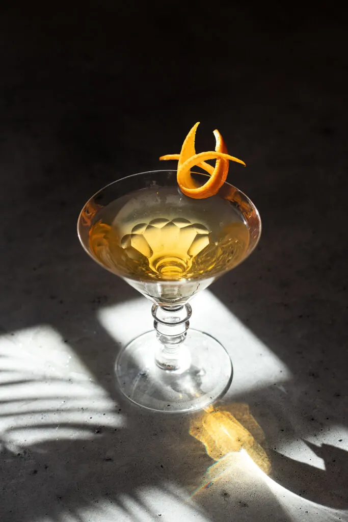 a golden colored cocktail in a martini glass with an orange twist.