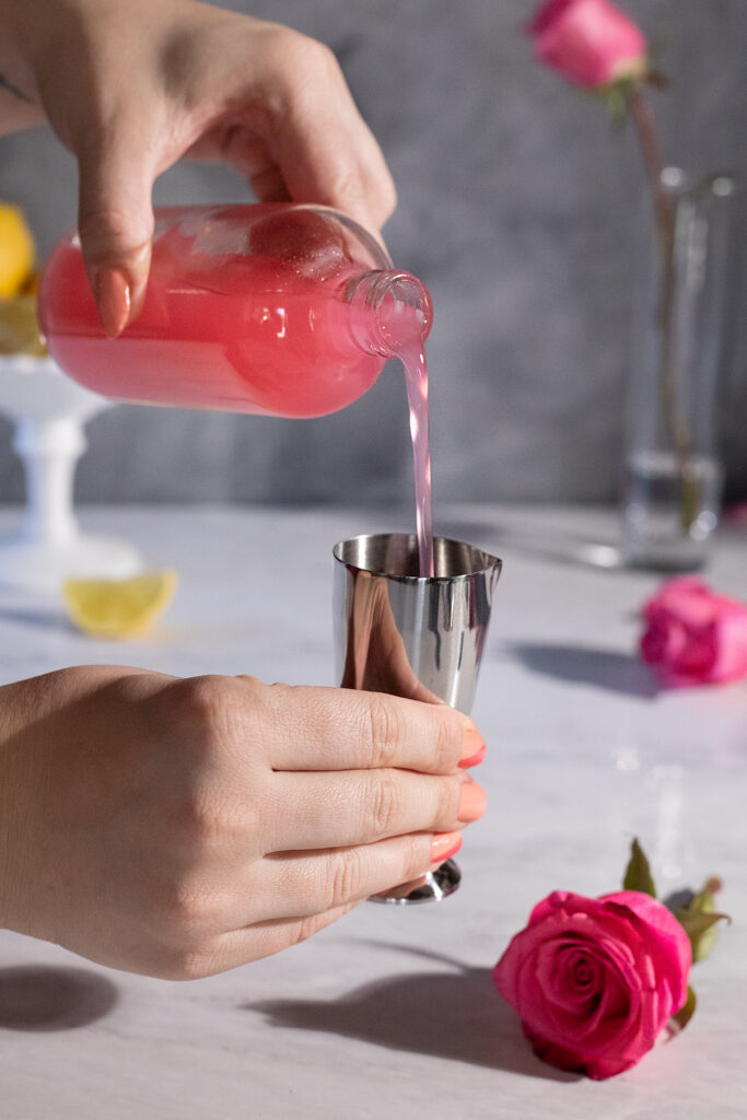 pouring pink syrup from bottle into a silver bar jigger next to roses.