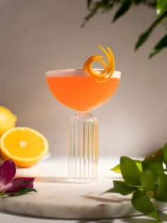 a bright orange liquid in an elegant coupe glass with a fancy orange twist.