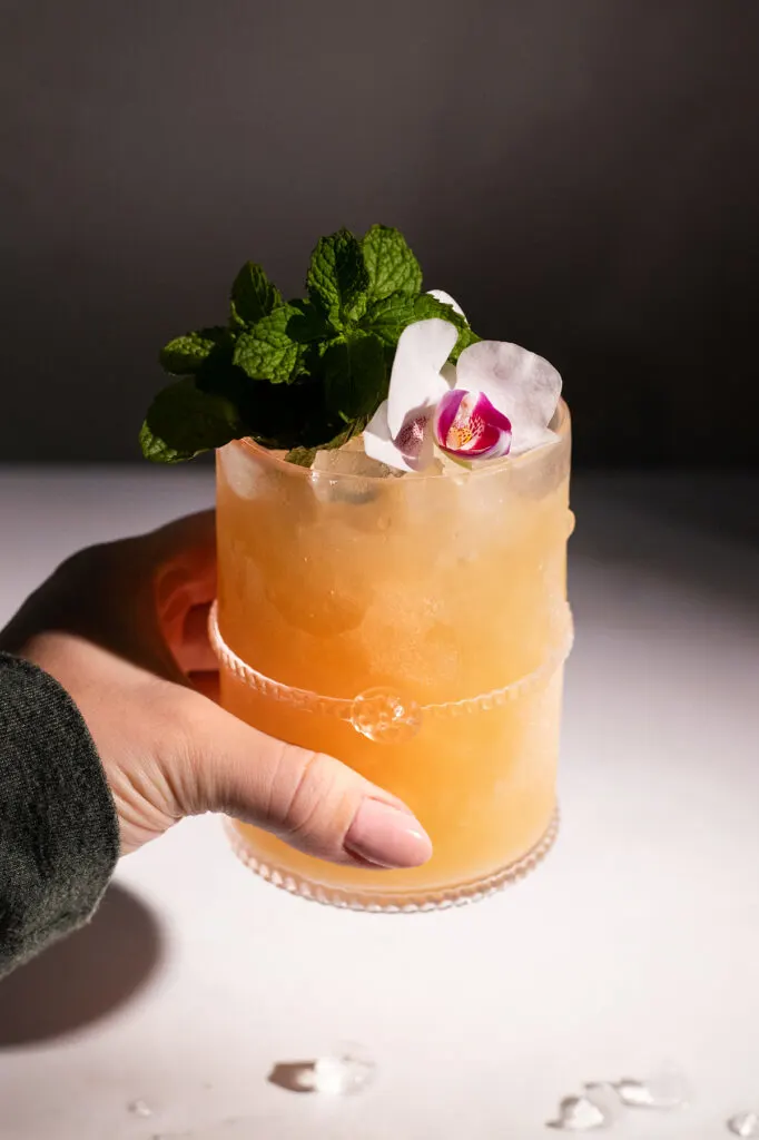 a woman's hand holding a tan colored cocktail in a rocks glass with mint and a flower.