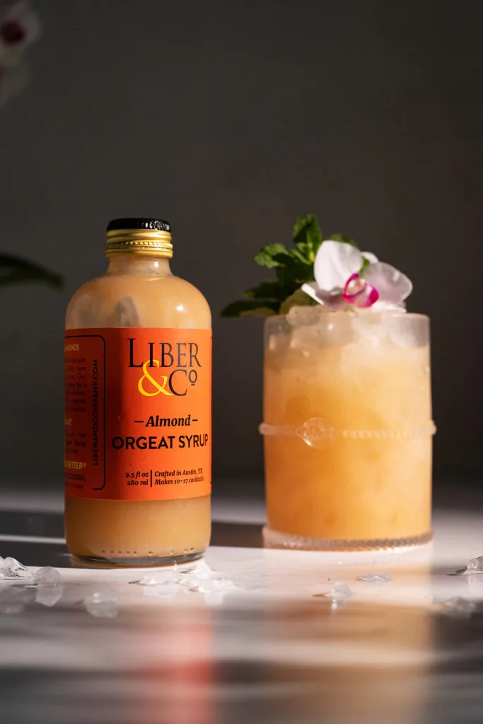 a bottle of tan Liber and Co almond orgeat syrup next to a cocktail.