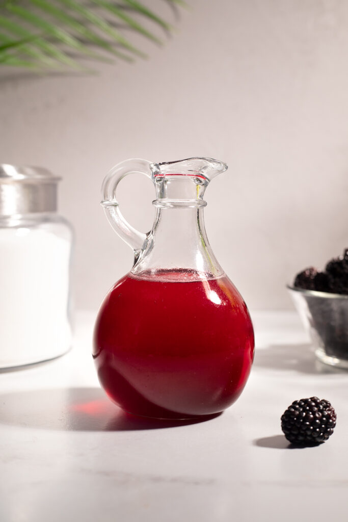 pitcher of dark red blackberry syrup on a white table.