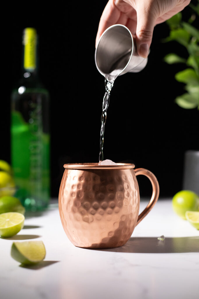 pouring clear cachaca from a metal jigger into a copper mule mug.