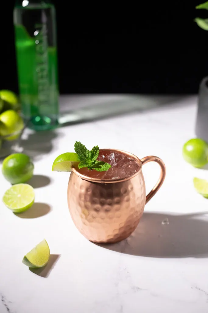copper mug filled with a Brazilian Buck cocktail next to limes and a bottle of Leblon Cachaca.