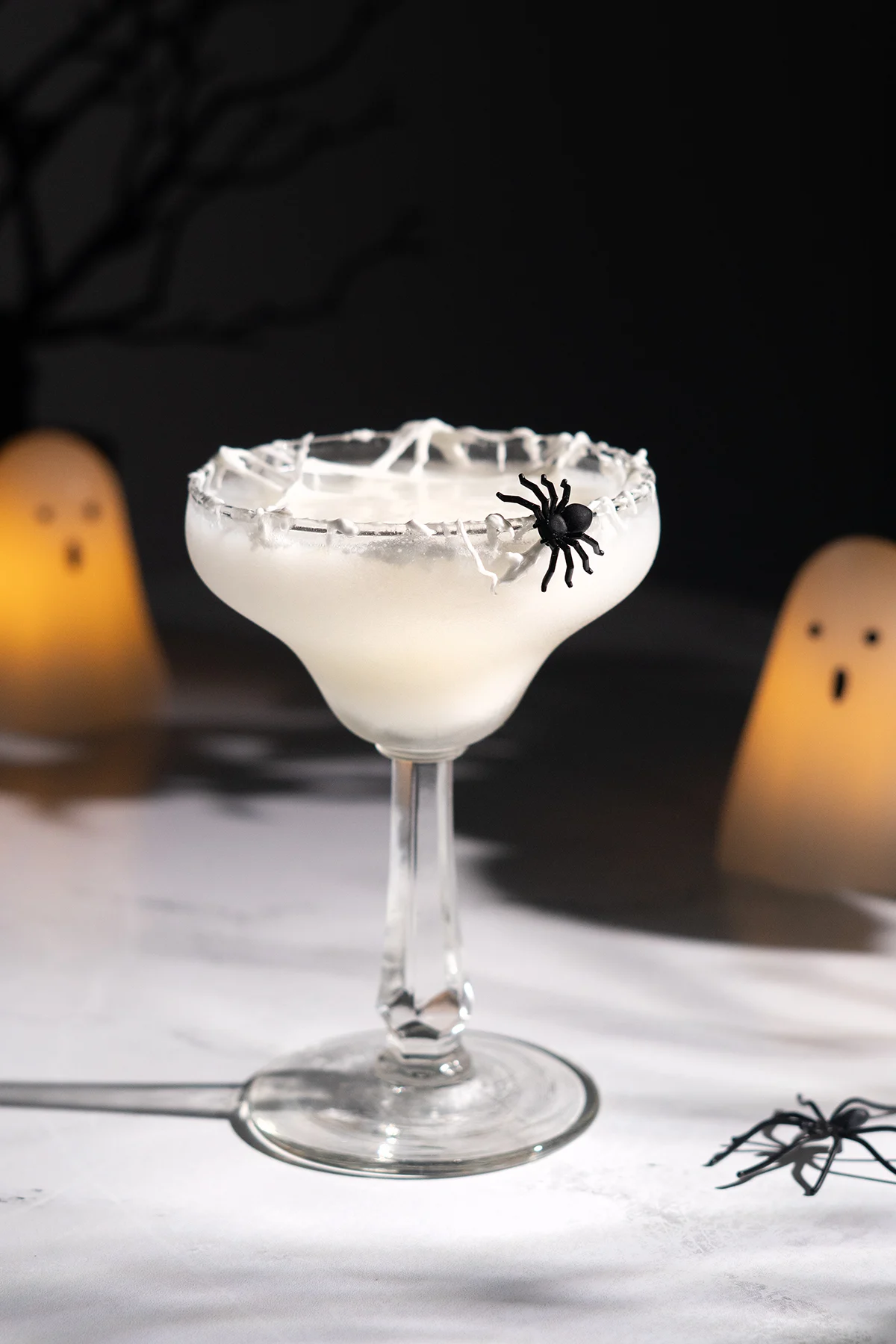 white cocktail with spider garnish next to ghost shaped candles.