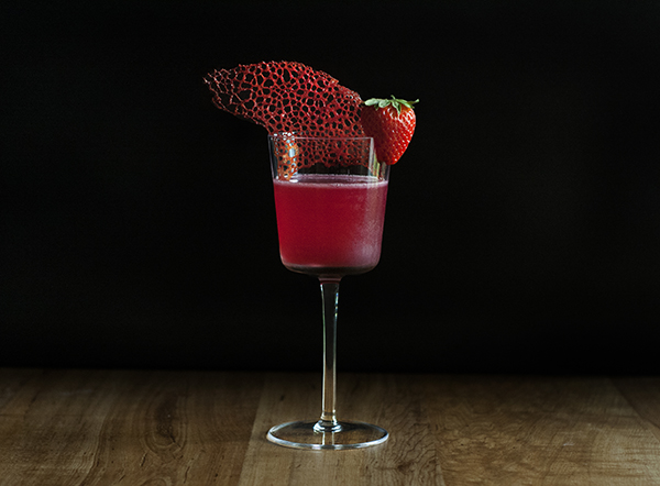 red cocktail garnished with a curved red coral tuile cookie and a strawberry.